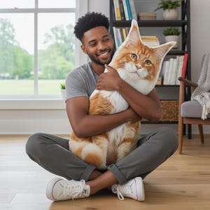 May include: A large, orange and white cat-shaped pillow is being hugged. The pillow has detailed facial features and a realistic fur pattern. The person holding the pillow is smiling. The background includes a bookshelf, window, and wooden floor.
