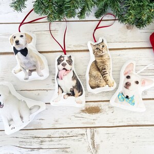 May include: Five white fabric ornaments with different animal faces printed on them. The ornaments are hanging from red ribbon and feature a dog wearing a bow tie, a cat, a dog with its tongue sticking out, a white dog, and a chihuahua wearing a blue bow tie.