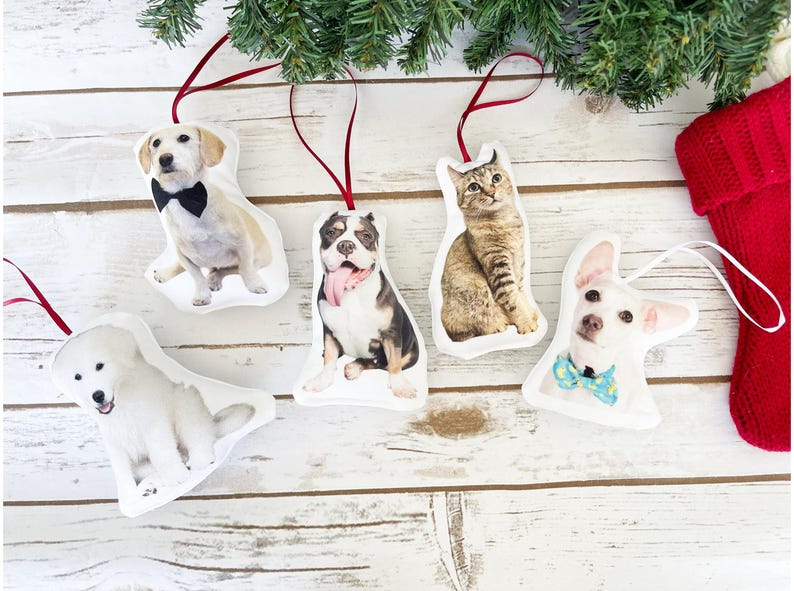 May include: Five white fabric ornaments with different animal faces printed on them. The ornaments are hanging from red ribbon and feature a dog wearing a bow tie, a cat, a dog with its tongue sticking out, a white dog, and a chihuahua wearing a blue bow tie.