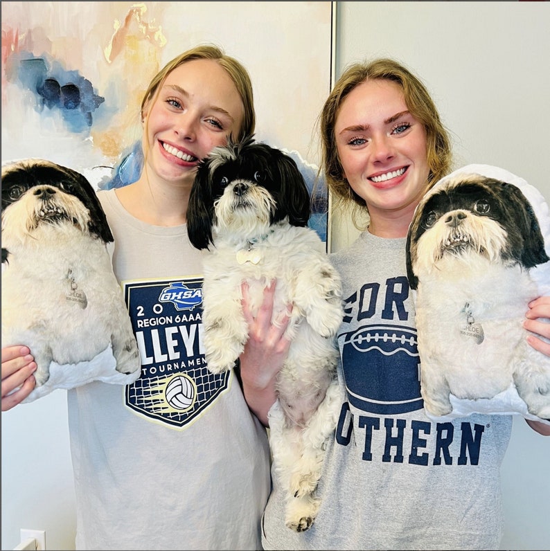 May include: Two young women are holding white fluffy dogs. The woman on the left is wearing a grey shirt with a volleyball tournament logo. The woman on the right is wearing a grey shirt with the text "Southern" in blue letters.