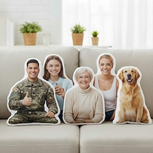 May include: A collection of custom-printed pillows shaped like people and a dog, arranged on a beige sofa. The pillows feature images of a man in military uniform, a young woman, an older woman, another young woman, and a golden retriever.