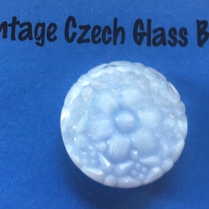 May include: A light blue, round Czech glass button with a floral design. The button has a raised, textured surface with a central flower and surrounding petals and leaves. The text "Vintage Czech Glass B" is above the button.