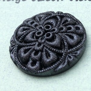 May include: A dark gray, oval-shaped button with an intricate floral design. The button has a raised, textured pattern with detailed swirls and leaf-like shapes. The button is likely made of metal or a similar material.