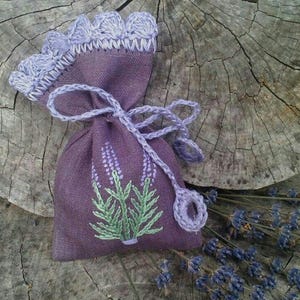 Può includere: Un sacchetto di lino viola con un disegno di lavanda e una parte superiore all'uncinetto. Il sacchetto è pieno di lavanda essiccata e si trova su una superficie di legno.
