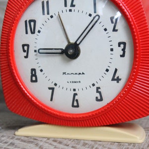 May include: A red alarm clock with a white face and black numbers. The clock has a white base and the words "Рясна" and "4 камня" printed on the face.