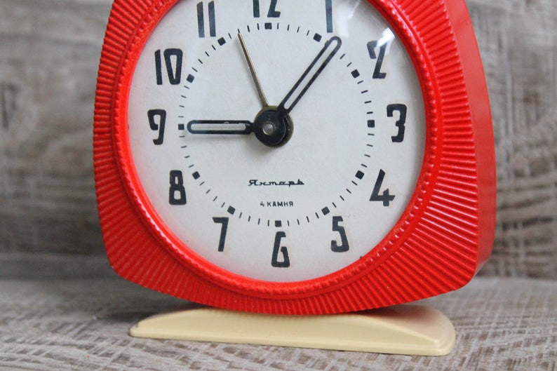 May include: A red alarm clock with a white face and black numbers. The clock has a white base and the words "Рясна" and "4 камня" printed on the face.