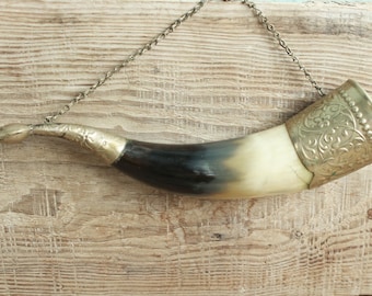 May include: A decorative drinking horn with a silver chain for hanging. The horn is a gradient of black to cream, with silver metal accents and ornate detailing around the rim. The horn is displayed against a rustic wooden background.