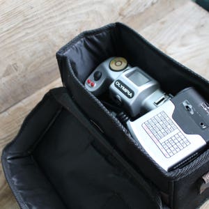 May include: A black camera bag with a silver camera and a black flash unit inside. The camera has the word "OLYMPIA" printed on it.