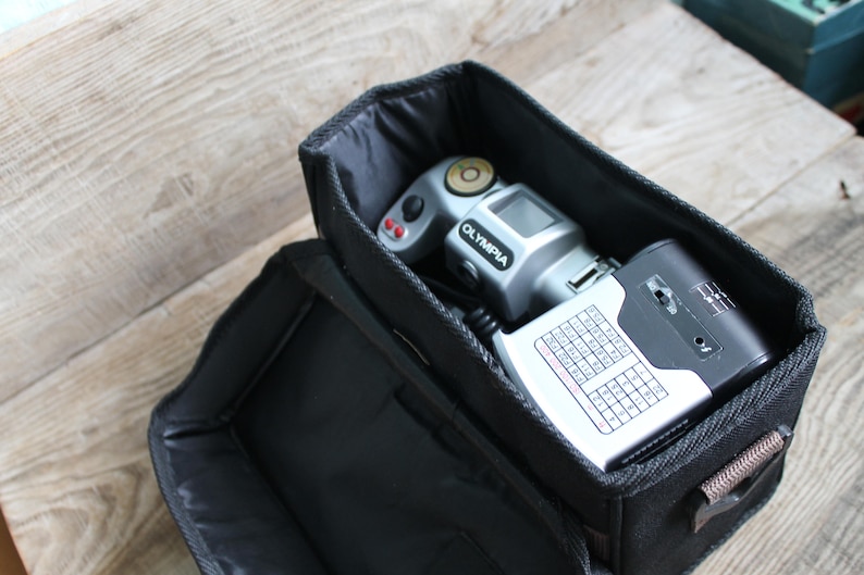 May include: A black camera bag with a silver camera and a black flash unit inside. The camera has the word "OLYMPIA" printed on it.