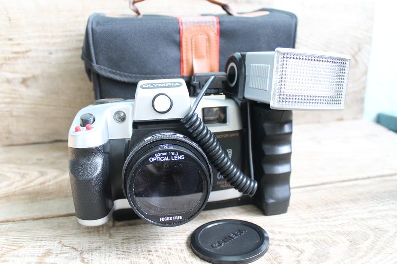 May include: A silver and black 35mm film camera with a black lens and a flash attachment. The camera has a red button and the words "OLYMPIA" and "50mm 1:8.3 OPTICAL LENS" printed on it.