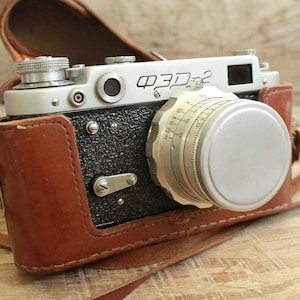 Puede incluir: Una cámara de película de 35 mm plateada y negra con una funda de cuero. La cámara tiene un objetivo con una tapa de lente plateada y una correa de cuero negra.