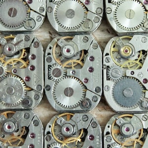 Puede incluir: Primer plano de varios movimientos de relojes plateados con engranajes intrincados y un vástago de cuerda de color dorado. Los movimientos de los relojes están dispuestos en filas sobre una superficie de madera. El texto "SU 1001-41" es visible en uno de los movimientos.
