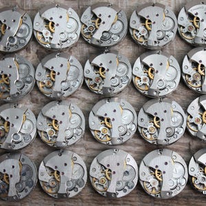 May include: A collection of 20 silver-toned watch movements with intricate gears and mechanisms. The watch movements are arranged in a grid pattern on a wooden surface.