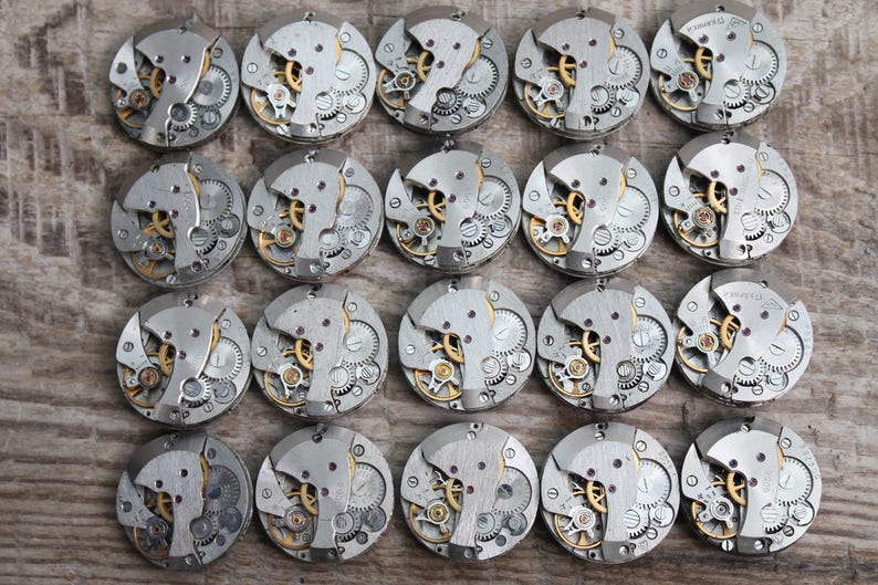 May include: A collection of 20 silver-toned watch movements with intricate gears and mechanisms. The watch movements are arranged in a grid pattern on a wooden surface.