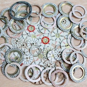 Puede incluir: Una colección de piezas de relojes antiguos, incluyendo ruedas de fecha y anillos giratorios, dispersas sobre una superficie de madera clara. Las piezas presentan detalles blancos, rojos y dorados con números y texto impresos, probablemente para reparación o creación de relojes.