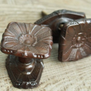 May include: Two square-shaped cabinet knobs with a dark brown finish. Each knob features a central circular element surrounded by radiating lines and a textured grid pattern on the four corners. The knobs have a short cylindrical stem and a base.