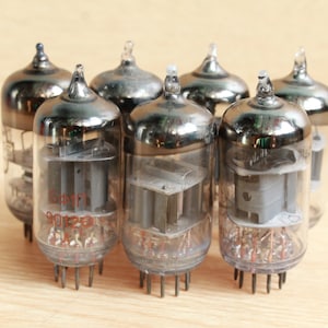 May include: A group of six vintage vacuum tubes with silver metal casings and glass bodies. The tubes are arranged in a circle on a light brown wooden surface.
