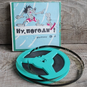 Puede incluir: Un juego de carrete y caja de película vintage. La caja presenta un lobo de dibujos animados persiguiendo a un conejo, con texto en ruso. La bobina es de color turquesa y contiene la tira de película. El conjunto está sobre una superficie de madera.