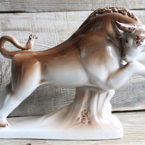 May include: A white and brown ceramic bull figurine with a raised tail and a small bird perched on its back. The bull is standing on a base with floral designs.