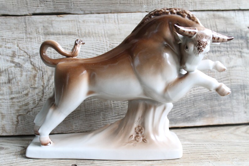 May include: A white and brown ceramic bull figurine with a raised tail and a small bird perched on its back. The bull is standing on a base with floral designs.