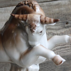 May include: A white porcelain bull figurine with brown accents. The bull is standing on its hind legs with its front legs raised. The bull has a detailed face and horns.