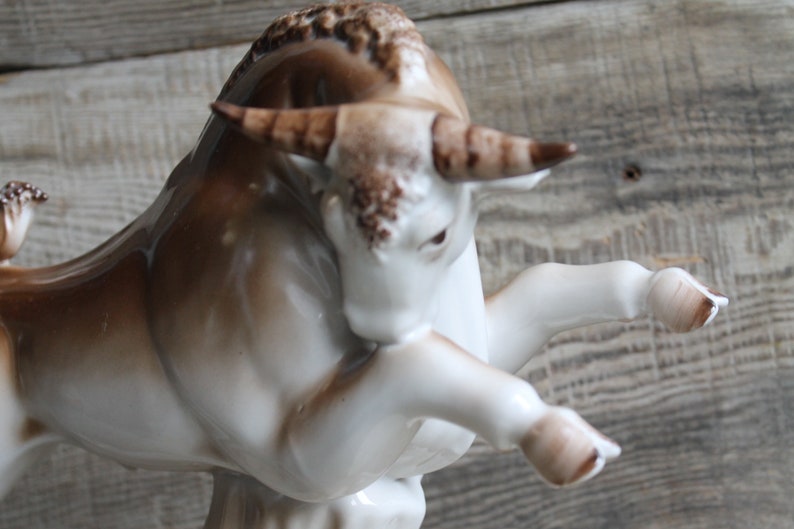 May include: A white porcelain bull figurine with brown accents. The bull is standing on its hind legs with its front legs raised. The bull has a detailed face and horns.