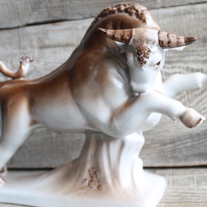 May include: A white porcelain bull figurine with brown accents. The bull is standing on its hind legs with its front legs raised. The figurine is on a white base with brown floral details.