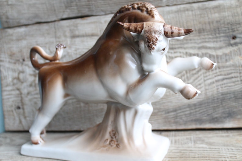 May include: A white porcelain bull figurine with brown accents. The bull is standing on its hind legs with its front legs raised. The figurine is on a white base with brown floral details.