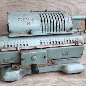 Puede incluir: Una calculadora mecánica vintage azul con el texto "Феликс М" en la parte superior. La calculadora tiene una serie de diales numerados y una palanca en el lateral. El número "188308" está impreso en la parte delantera.
