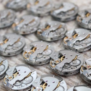 May include: Close-up of multiple silver watch movements with intricate gears and mechanisms. The watch parts are arranged in rows on a light wood surface.
