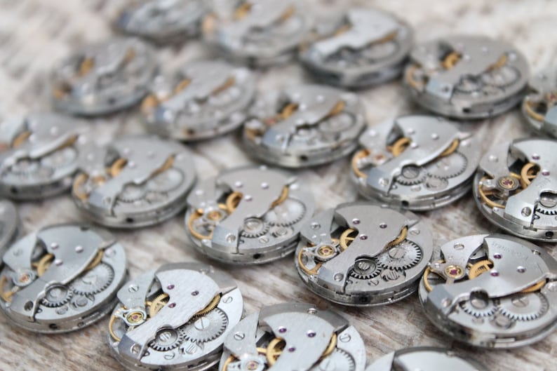 May include: Close-up of multiple silver watch movements with intricate gears and mechanisms. The watch parts are arranged in rows on a light wood surface.