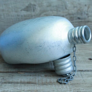 May include: A silver-colored canteen with a screw-on cap and a chain. The canteen has a rounded body and a textured surface. The cap is attached to the canteen with a small chain.