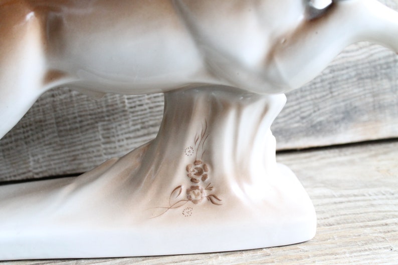 May include: White porcelain figurine of a deer with brown accents. The deer is in a running pose with its head turned to the side. The base of the figurine is decorated with a brown floral design.