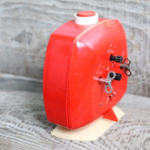 May include: A red wind-up alarm clock with a white base and two wind-up keys. The clock has a square face and a small red button on top.