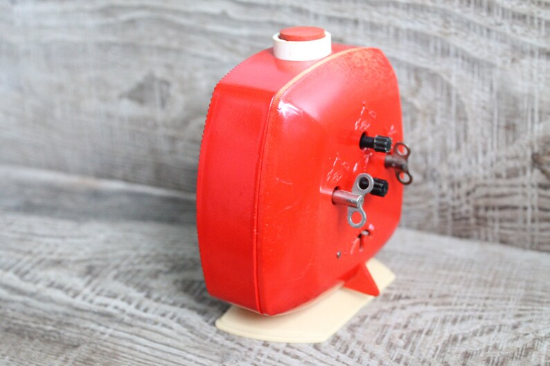 May include: A red wind-up alarm clock with a white base and two wind-up keys. The clock has a square face and a small red button on top.