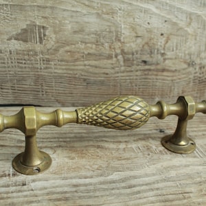 Puede incluir: Manija de puerta de latón antiguo con un agarre central en forma de piña. El mango tiene un diseño detallado y texturizado y está montado sobre una superficie de madera. La ferretería es de color dorado cálido.
