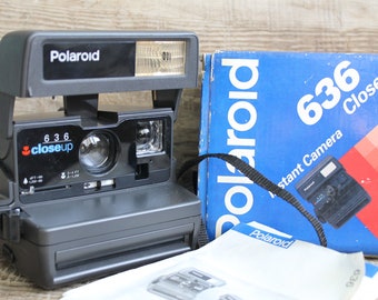 May include: A gray Polaroid 636 CloseUp instant camera with a built-in flash, next to its blue box. The box has the Polaroid logo and the camera model name. The camera has a black strap and a manual.