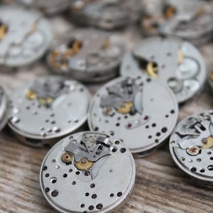 May include: Close-up of several silver watch movements with intricate gears and mechanisms. The watch parts are arranged on a wooden surface.