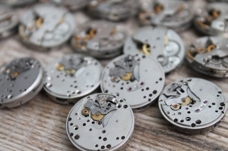 May include: Close-up of several silver watch movements with intricate gears and mechanisms. The watch parts are arranged on a wooden surface.