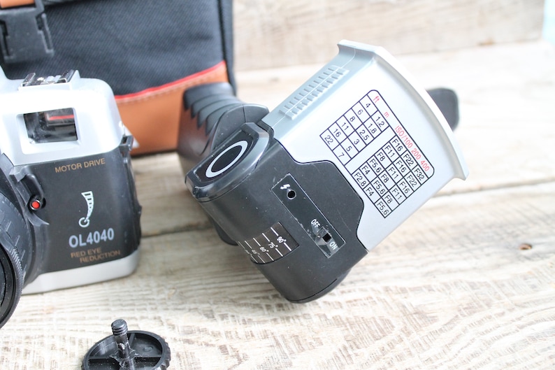 May include: A black and silver camera with a motor drive and red eye reduction. The camera has a flash unit attached to it. The flash unit has a chart with ISO and aperture settings.