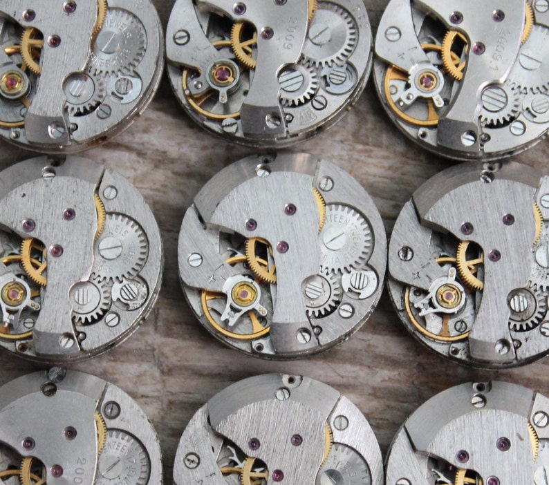 May include: Close-up of ten silver watch movements with intricate gears and a gold finish. The watch movements are arranged in a grid pattern on a wooden surface.
