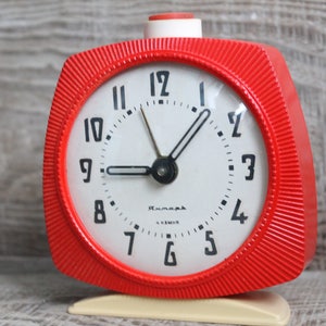 May include: A red vintage alarm clock with a white face and black hands. The clock has a white base and a red button on the top. The clock face has black numbers and the word "Rumania" printed on it.