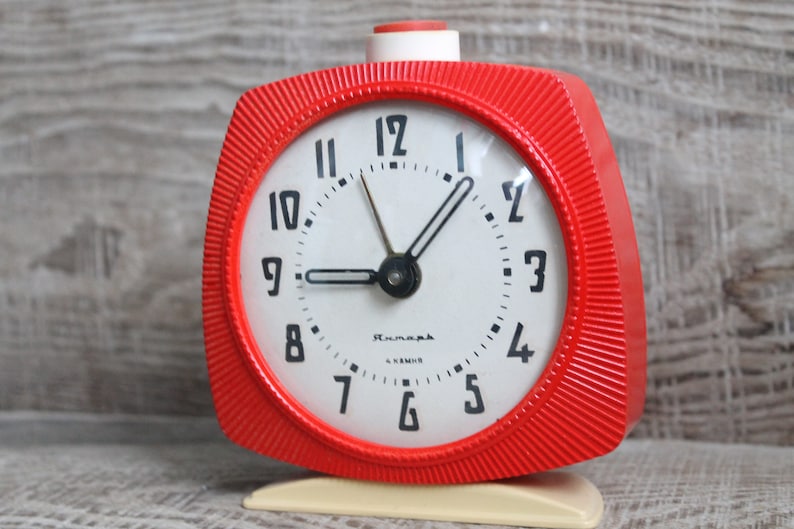 May include: A red vintage alarm clock with a white face and black hands. The clock has a white base and a red button on the top. The clock face has black numbers and the word "Rumania" printed on it.