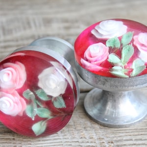 Op de afbeelding: Twee decoratieve kastknoppen met een rode achtergrond en een bloemenontwerp. Elke knop heeft een heldere, afgeronde bovenkant met witte en roze rozen en groene bladeren. De knoppen hebben een zilverkleurige basis.