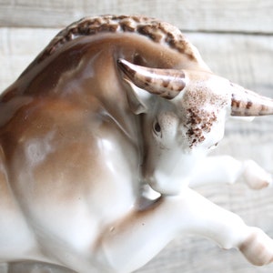 May include: A white porcelain bull figurine with brown accents. The bull is in a charging pose with its head lowered and horns pointed forward.