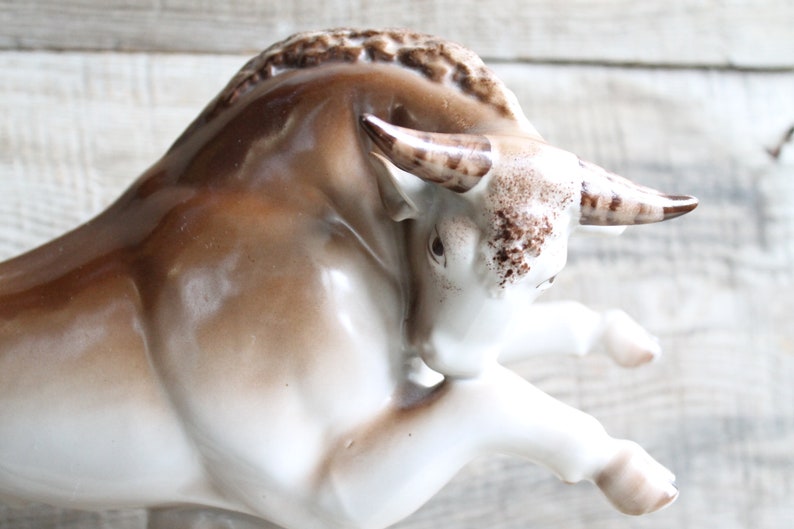 May include: A white porcelain bull figurine with brown accents. The bull is in a charging pose with its head lowered and horns pointed forward.