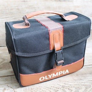 May include: A black canvas camera bag with brown leather accents and a brown leather strap. The bag has a black buckle and the word "OLYMPIA" is printed in white on the front.