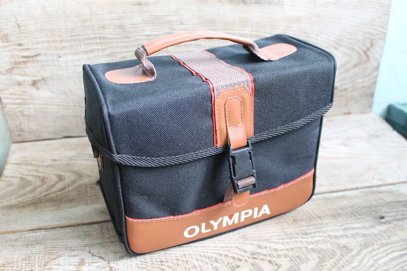 May include: A black canvas camera bag with brown leather accents and a brown leather strap. The bag has a black buckle and the word "OLYMPIA" is printed in white on the front.