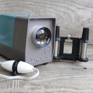 May include: A vintage blue and gray slide projector with a black slide tray and a white cord. The projector has a lens with a black rim and the word "ЭКРАН" printed on the front.