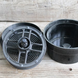 May include: A black film developing tank with a spiral reel and a lid, set against a light wooden surface. The tank is open, revealing the interior components used for processing photographic film.
